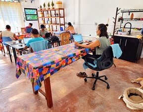 Spacious coworking area with several people working on laptops at colorful communal tables, assorted chairs, wooden display shelving, kitchenette, and a dog resting on the floor.