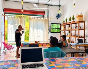 Colorful coworking area with people working and talking around communal tables, laptops, bright woven chairs, hanging lights, wooden shelving, and large windows.