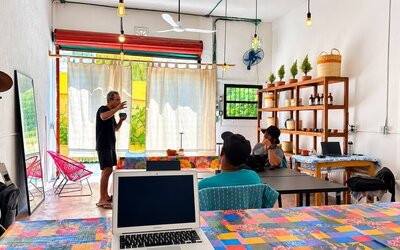 Colorful coworking area with people working and talking around communal tables, laptops, bright woven chairs, hanging lights, wooden shelving, and large windows.