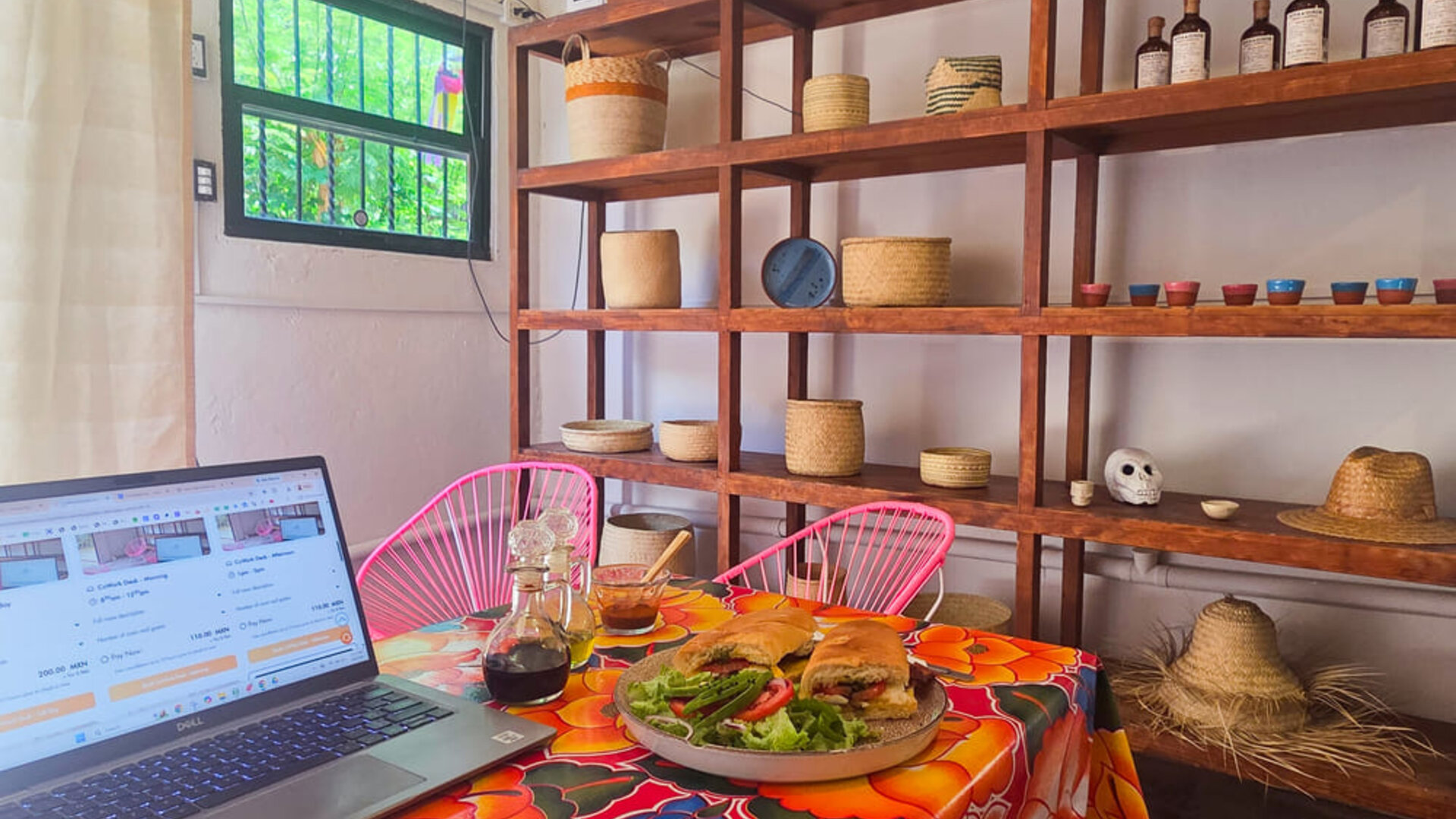 Laptop beside a plated sandwich and fresh salad on a colorful floral tablecloth, with pink chairs and wooden shelving displaying woven baskets and decorative items.