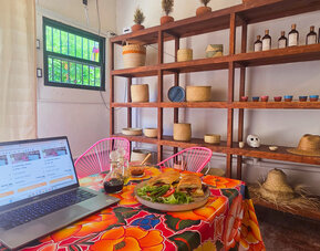 Laptop beside a plated sandwich and fresh salad on a colorful floral tablecloth, with pink chairs and wooden shelving displaying woven baskets and decorative items.