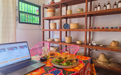 Laptop beside a plated sandwich and fresh salad on a colorful floral tablecloth, with pink chairs and wooden shelving displaying woven baskets and decorative items.