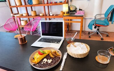 Open laptop beside a plated breakfast of omelet, black beans, potatoes, and cheese on a spacious coworking table with colorful seating and decorative shelving.