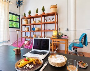Bright coworking area with an open laptop and breakfast on a large black table, surrounded by pink chairs, wooden shelving, woven décor, plants, and warm pendant lighting.