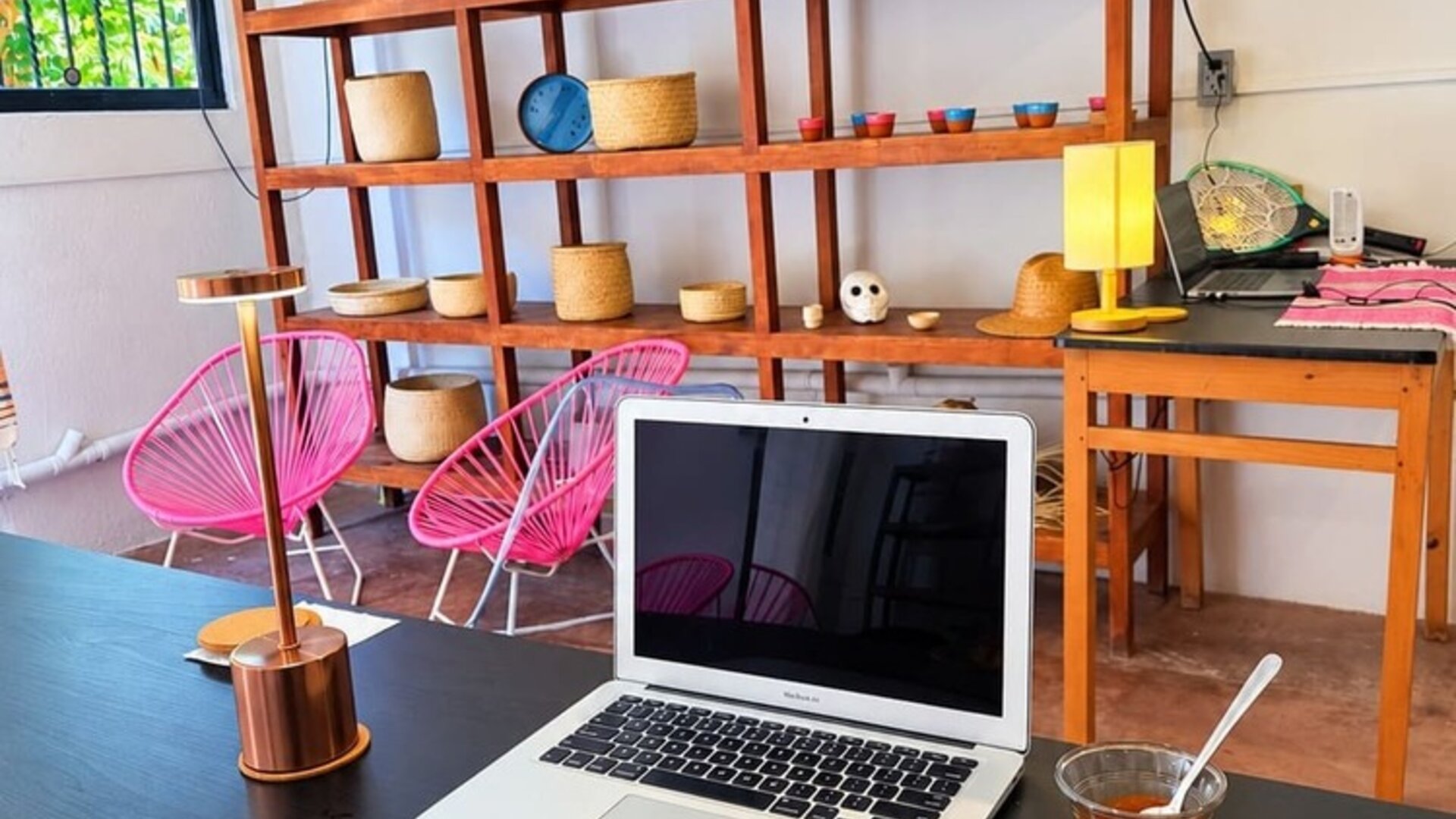 Breakfast plate with an omelet, black beans, potatoes, and fresh garnishes beside an open laptop in a bright coworking space with pink chairs and wooden shelving.