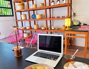 Breakfast plate with an omelet, black beans, potatoes, and fresh garnishes beside an open laptop in a bright coworking space with pink chairs and wooden shelving.