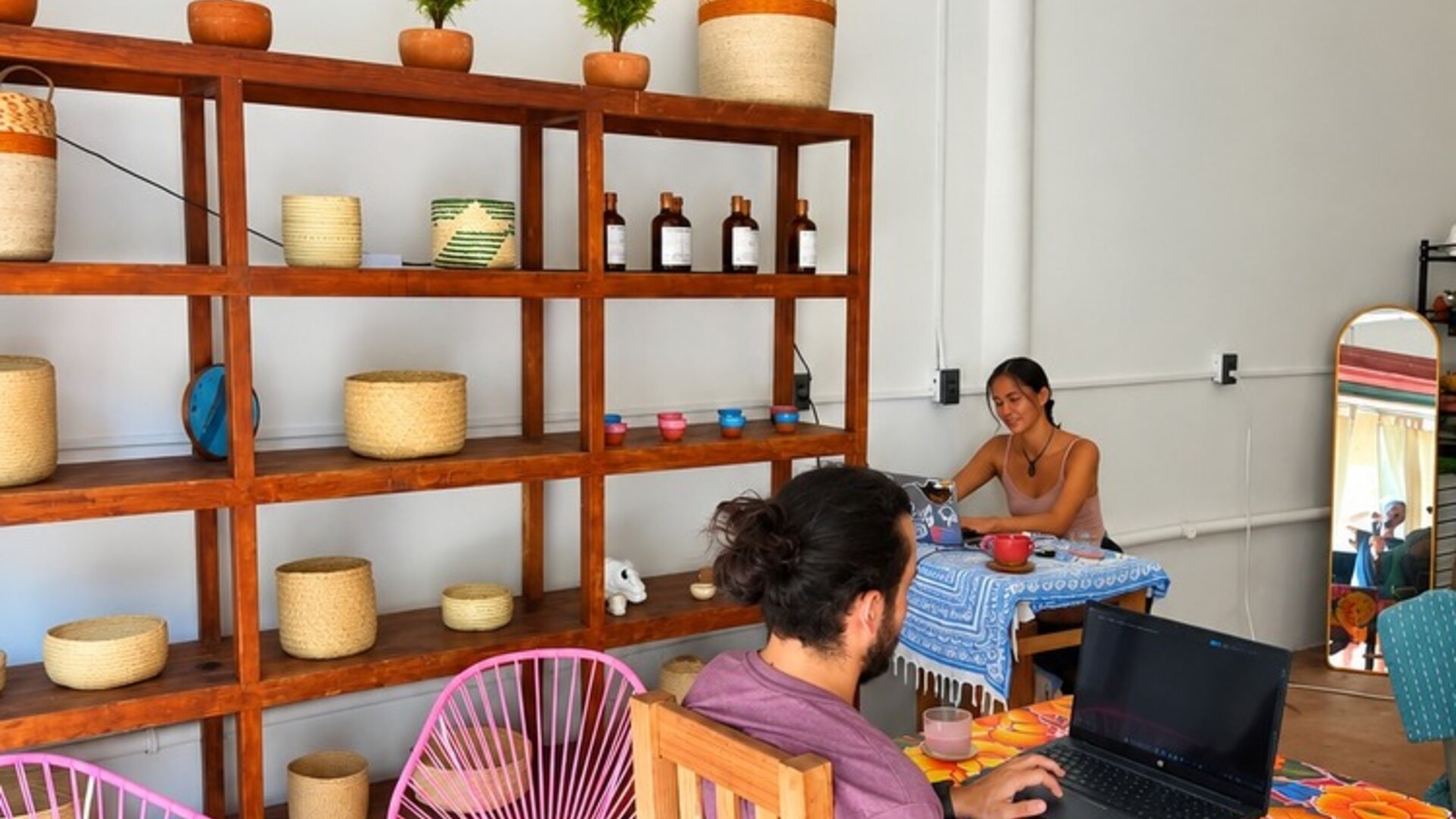 Relaxed coworking space with people working on laptops at colorful tables, pink woven chairs, decorative wooden shelving, potted plants, and warm pendant lighting.