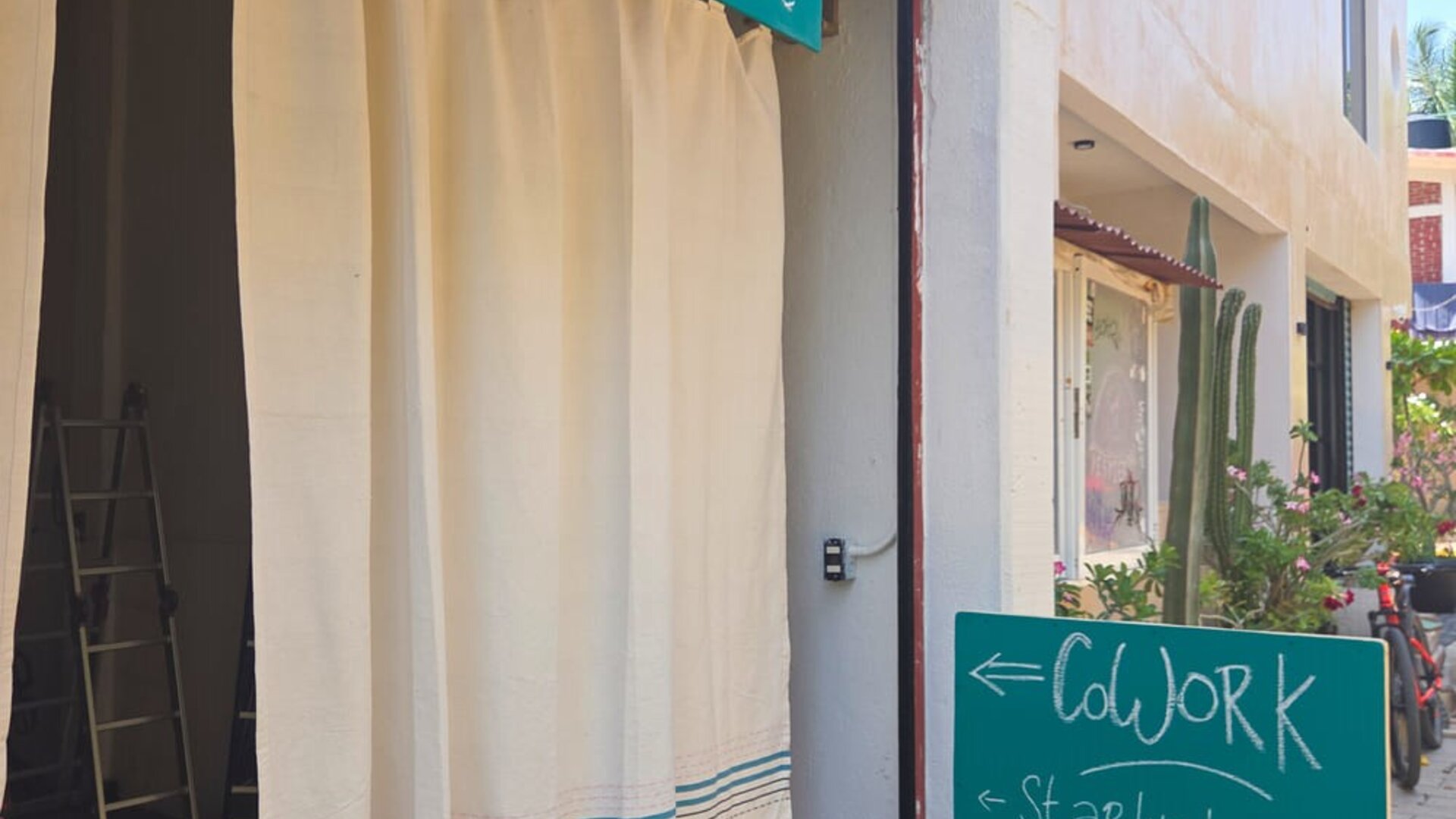 Entrance to a zen coworking space with white curtains and turquoise signs advertising Wi-Fi, office chairs, café food, and a half-day coworking rate.