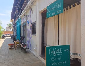 Street view of a relaxed coworking space entrance with turquoise signage, white curtains, parked motorcycles, and neighboring buildings along a sunny pedestrian lane.
