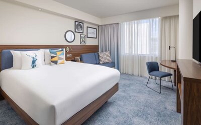 Bright contemporary king room with a white bed, blue upholstered headboard, colorful pillows, sofa, work desk, chair, large windows and wall-mounted TV.