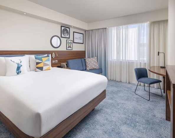 Bright contemporary king room with a white bed, blue upholstered headboard, colorful pillows, sofa, work desk, chair, large windows and wall-mounted TV.