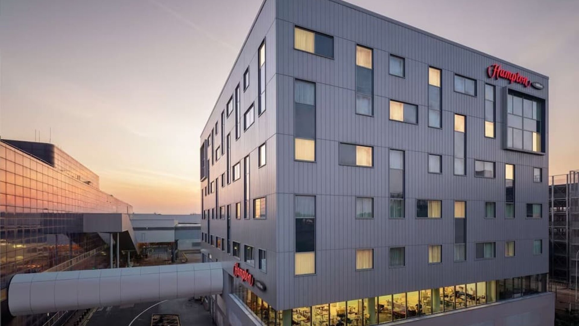 Modern Hampton hotel exterior at sunset with a gray multi-story façade, illuminated guest room windows and an enclosed walkway connecting to a neighboring building.