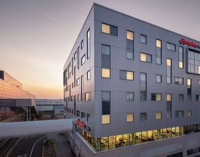 Modern Hampton hotel exterior at sunset with a gray multi-story façade, illuminated guest room windows and an enclosed walkway connecting to a neighboring building.