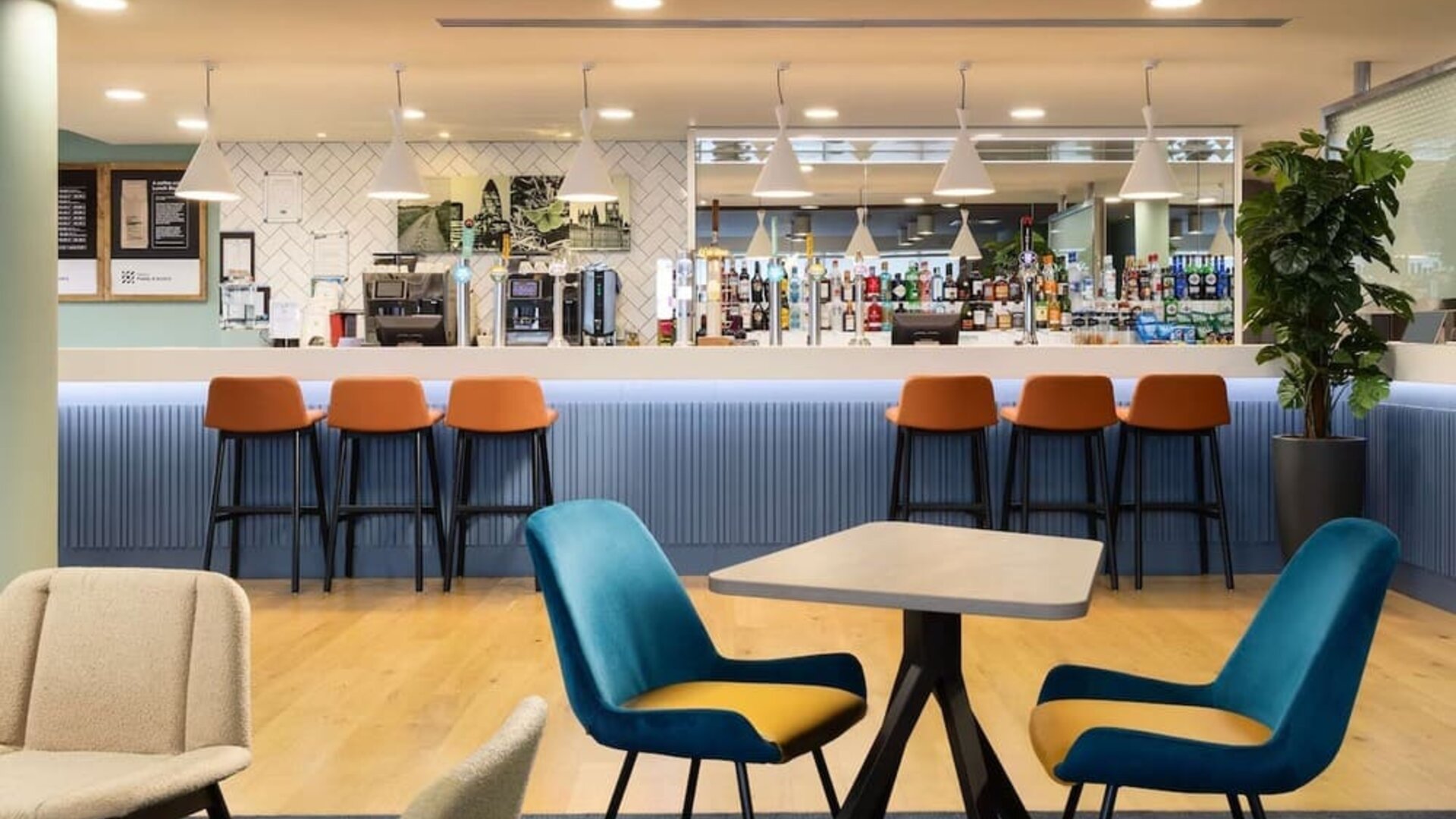 Contemporary hotel bar and lounge with a long blue counter, orange bar stools, pendant lighting, stocked drinks display, wooden flooring and blue lounge chairs.