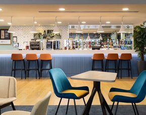 Contemporary hotel bar and lounge with a long blue counter, orange bar stools, pendant lighting, stocked drinks display, wooden flooring and blue lounge chairs.