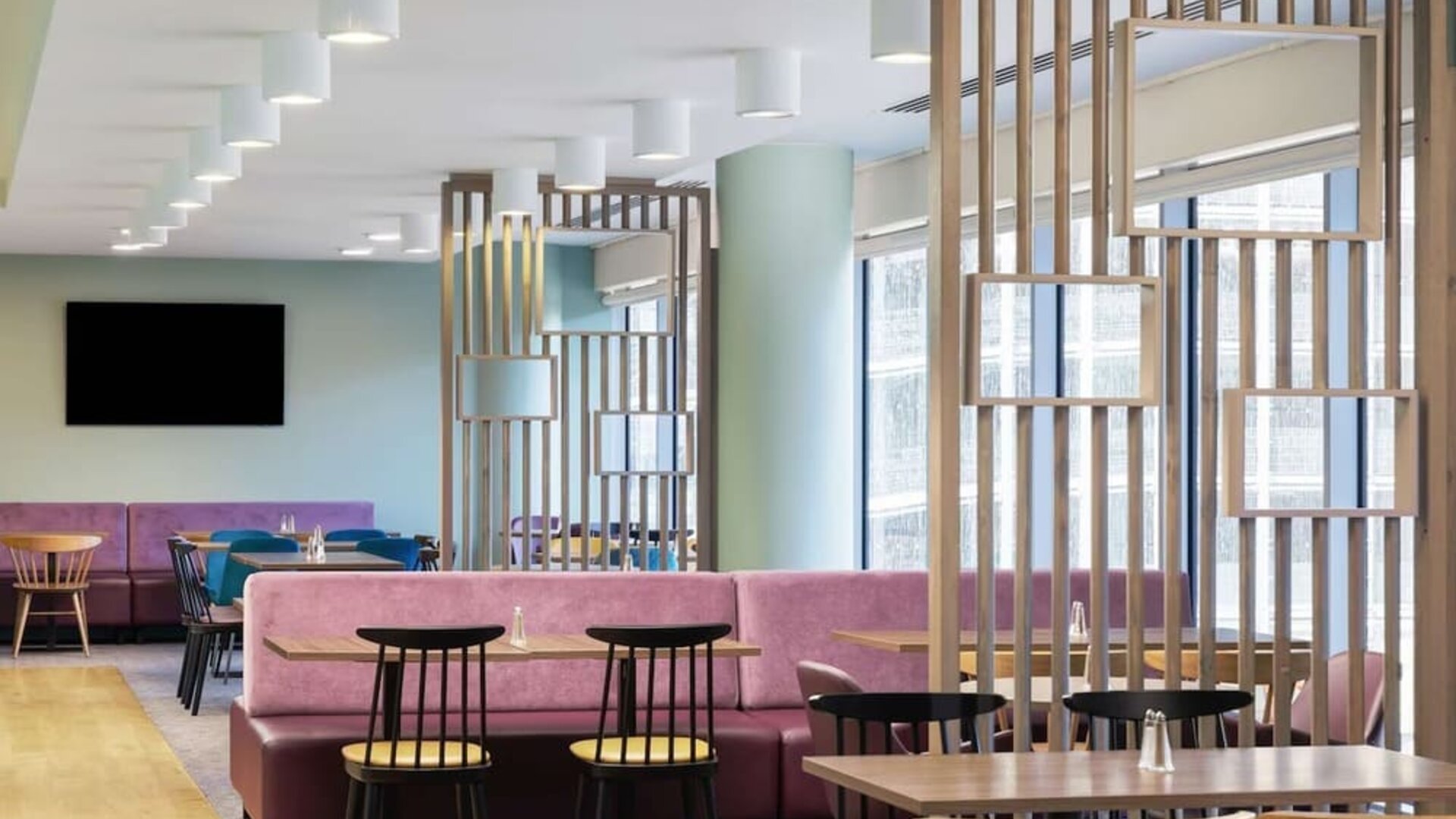 Modern hotel dining and coworking area with pink upholstered banquette seating, wooden tables, black chairs, decorative wood partitions, mirrors and large windows.