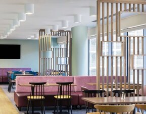 Modern hotel dining and coworking area with pink upholstered banquette seating, wooden tables, black chairs, decorative wood partitions, mirrors and large windows.