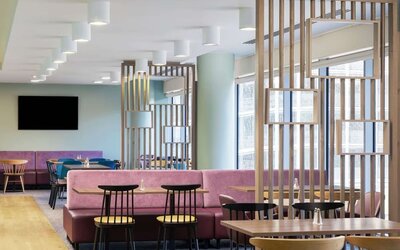 Modern hotel dining and coworking area with pink upholstered banquette seating, wooden tables, black chairs, decorative wood partitions, mirrors and large windows.