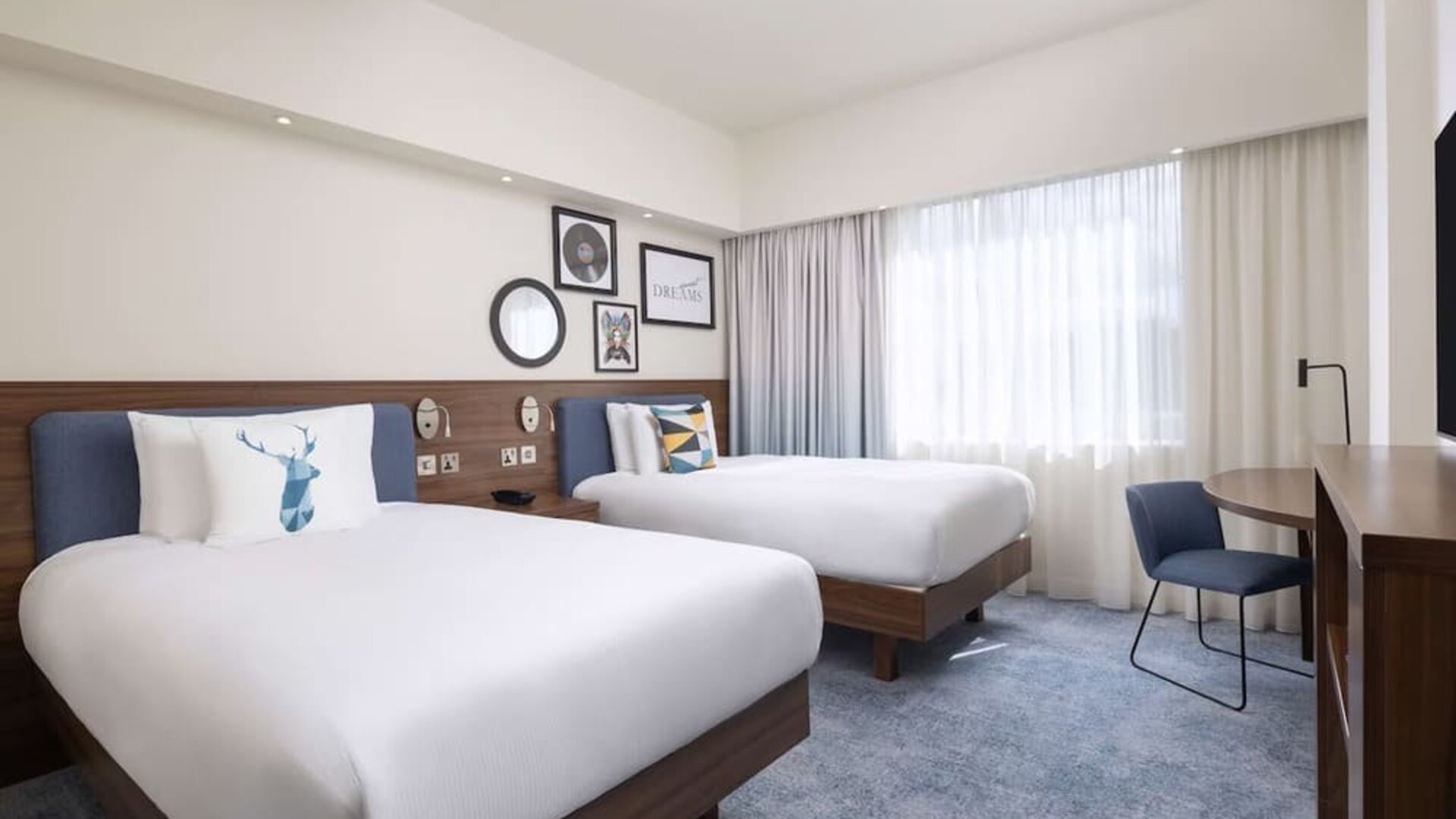 Spacious hotel twin room with two white beds, blue upholstered headboards, decorative pillows, large curtained windows, work desk, chair and wooden TV console.