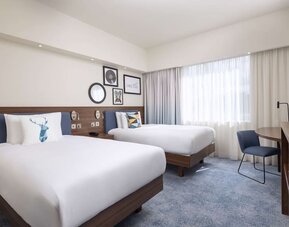 Spacious hotel twin room with two white beds, blue upholstered headboards, decorative pillows, large curtained windows, work desk, chair and wooden TV console.