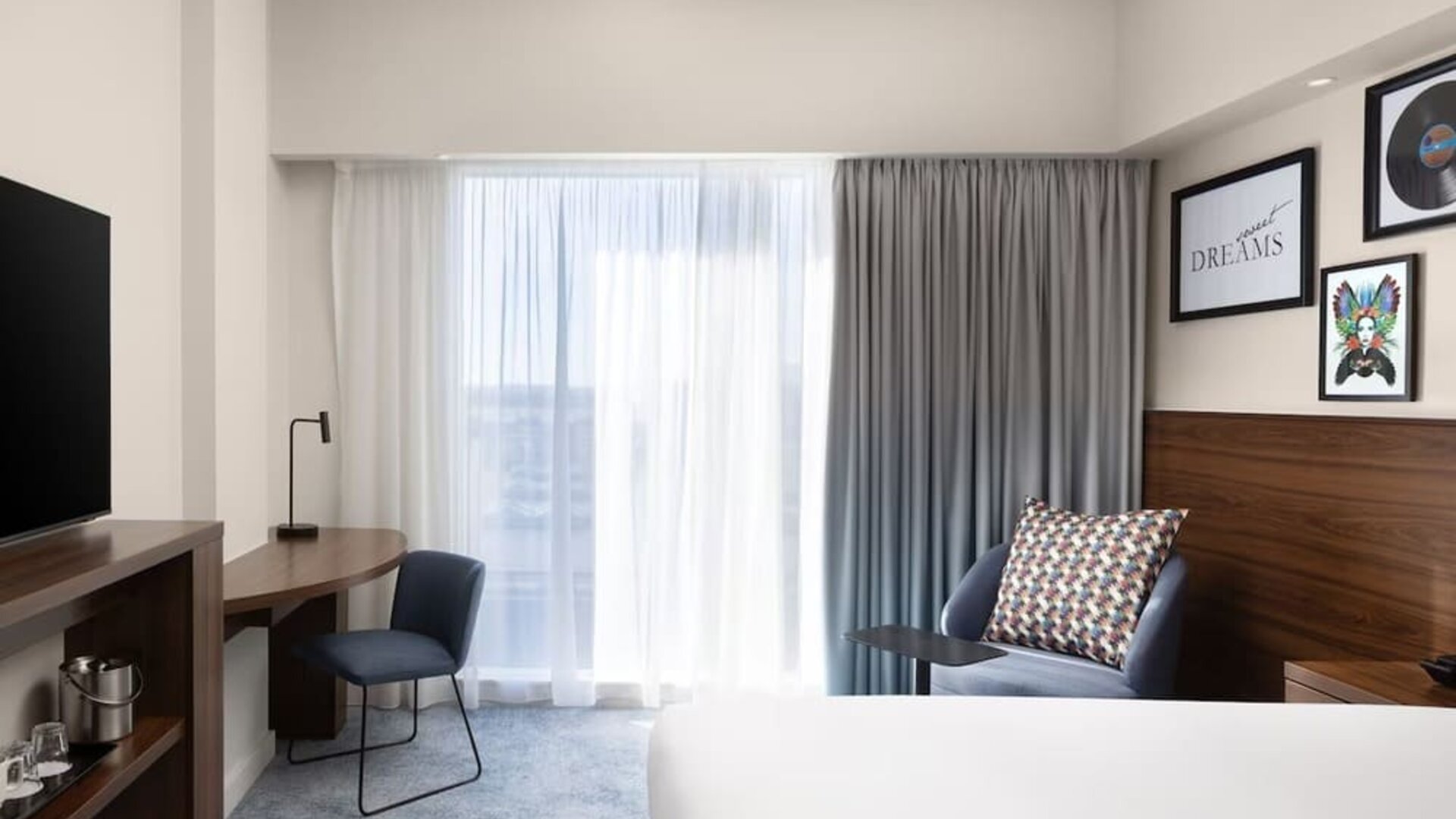 Bright modern hotel room with a large bed, floor-to-ceiling window and curtains, blue armchair, wooden work desk, chair, wall-mounted TV and contemporary artwork.