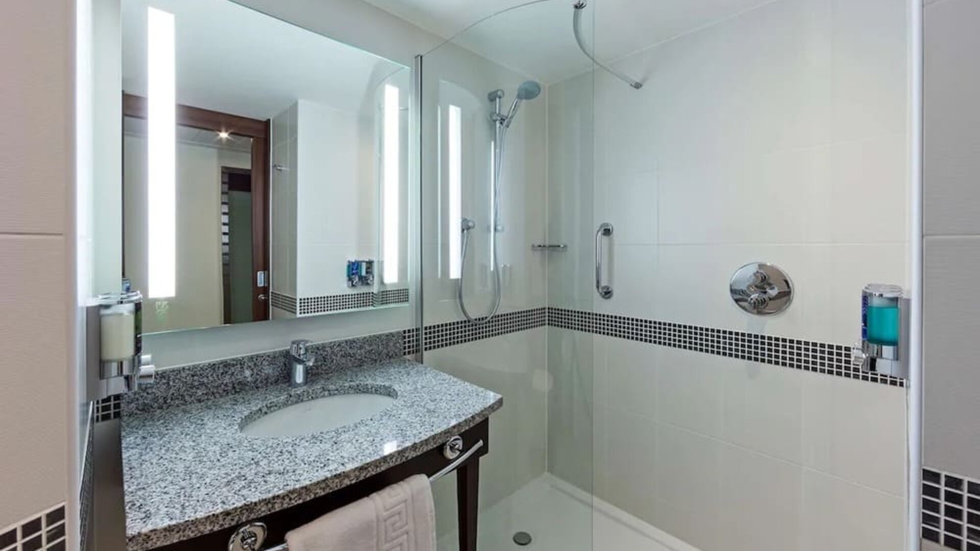 Modern hotel bathroom with a granite vanity, illuminated mirror, glass-enclosed walk-in shower, handheld showerhead, grab rail and wall-mounted toiletries.