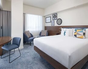 Spacious modern king room with a large white bed, blue upholstered headboard, colorful decorative pillows, two armchairs, work desk and large curtained windows.