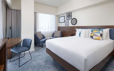 Spacious modern king room with a large white bed, blue upholstered headboard, colorful decorative pillows, two armchairs, work desk and large curtained windows.