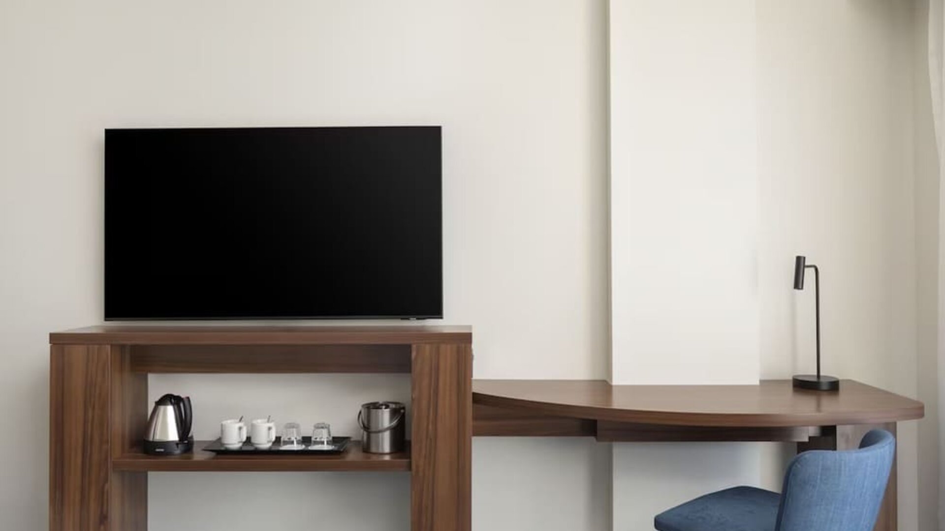 Hotel room workspace with a large flat-screen TV, curved wooden desk, blue chair, desk lamp and hospitality station with kettle, cups, glasses and ice bucket.