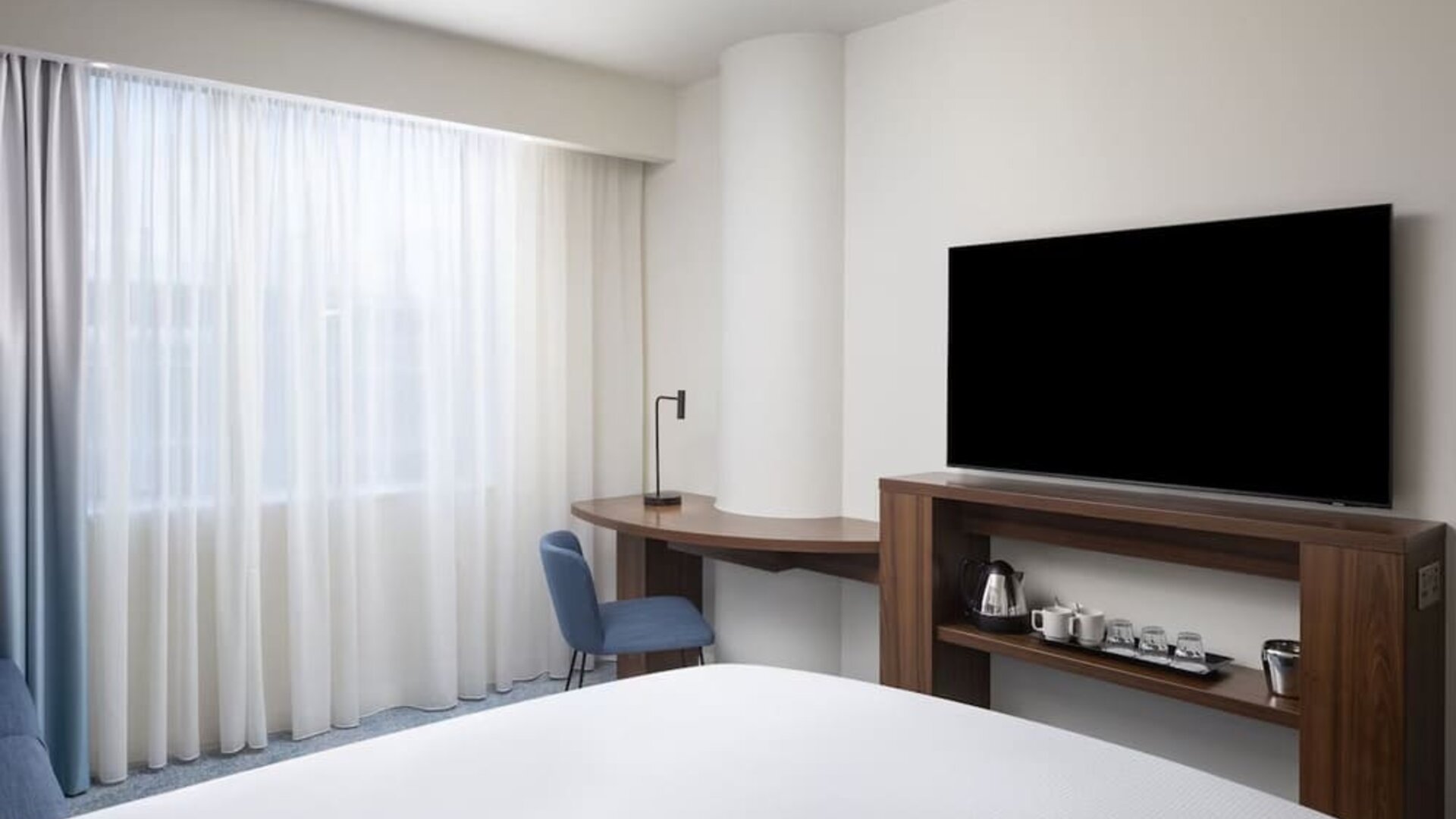 Bright contemporary hotel room with a large bed, floor-to-ceiling sheer curtains, wooden work desk, blue chair, wall-mounted TV and tea and coffee station.