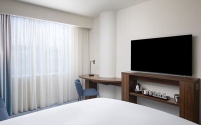 Bright contemporary hotel room with a large bed, floor-to-ceiling sheer curtains, wooden work desk, blue chair, wall-mounted TV and tea and coffee station.