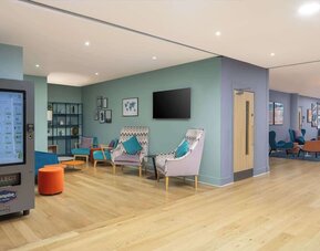 Colorful contemporary hotel lobby with teal and patterned lounge chairs, small tables, pale wood flooring, pastel green and blue walls and multiple seating areas.