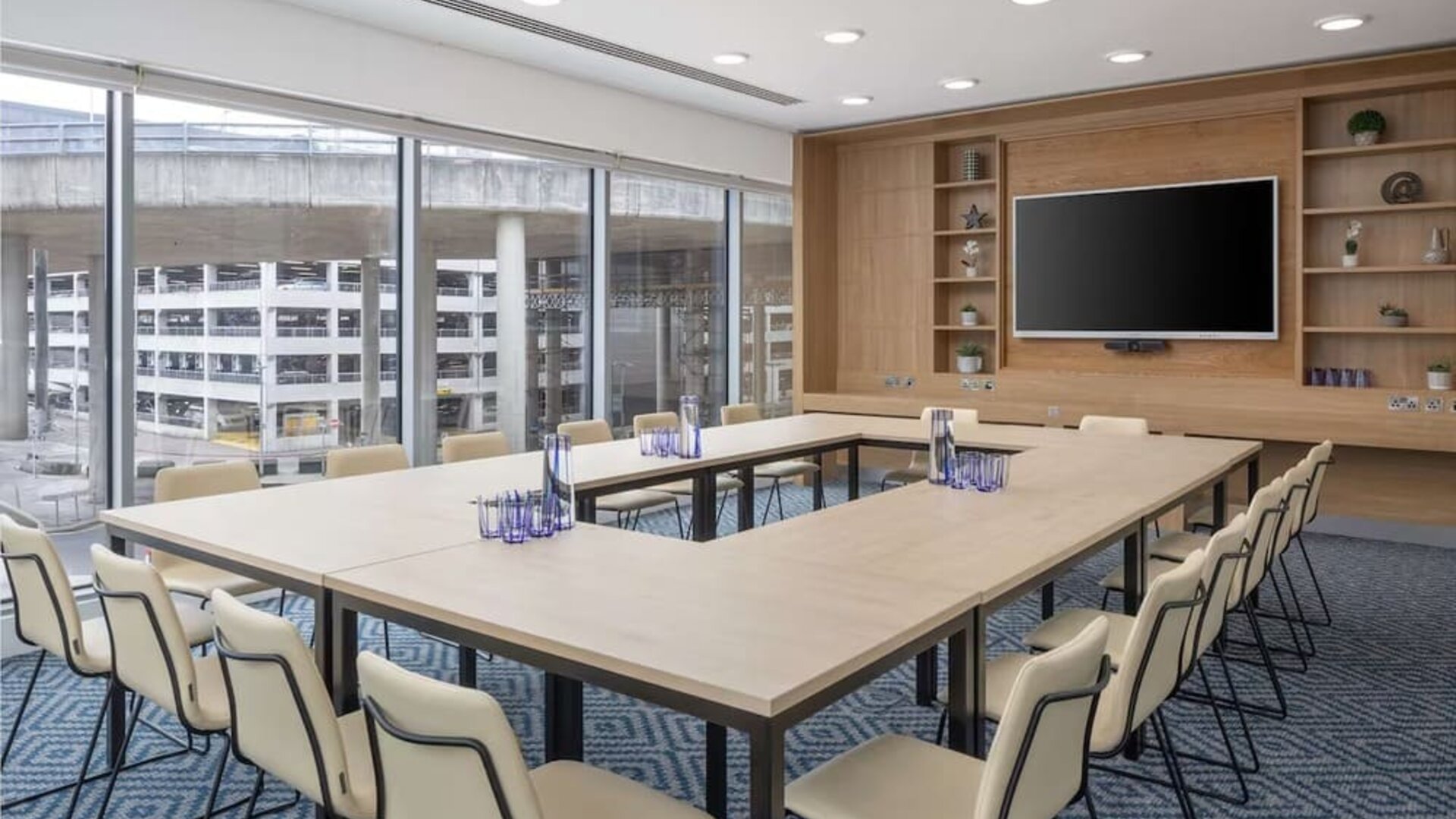 Bright hotel meeting room with U-shaped conference tables, upholstered chairs, large wall-mounted TV, wooden shelving and floor-to-ceiling windows overlooking nearby buildings.