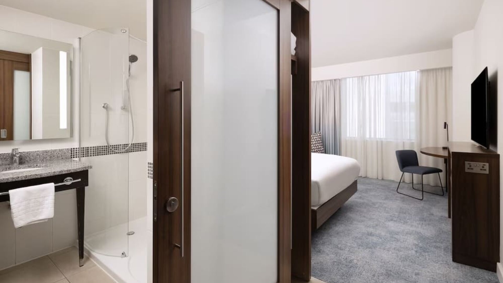 Contemporary hotel room viewed from the entrance, with a king bed, work desk and TV beside a modern bathroom with a glass-enclosed shower and illuminated mirror.