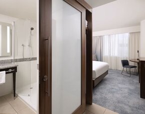 Contemporary hotel room viewed from the entrance, with a king bed, work desk and TV beside a modern bathroom with a glass-enclosed shower and illuminated mirror.