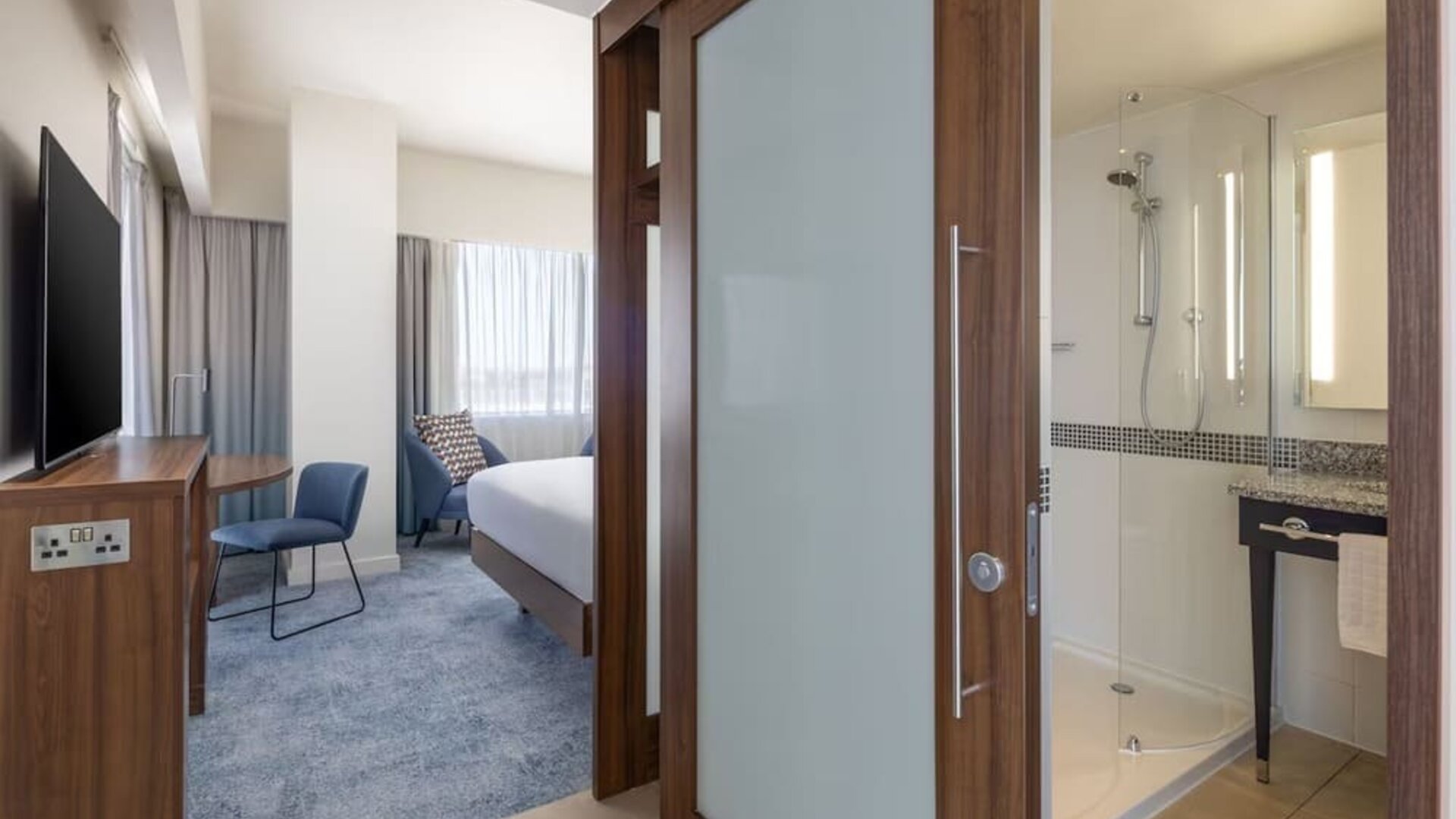 Modern hotel king suite with a bedroom and bathroom separated by a frosted-glass sliding door, featuring a work desk, wall-mounted TV and glass-enclosed shower.