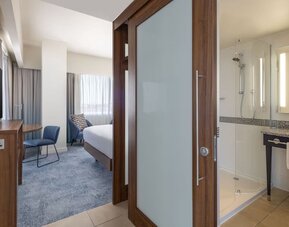 Modern hotel king suite with a bedroom and bathroom separated by a frosted-glass sliding door, featuring a work desk, wall-mounted TV and glass-enclosed shower.