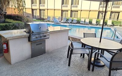 Outdoor hotel poolside terrace with built-in stainless steel grill, round dining table, chairs and large umbrella overlooking the swimming pool and loungers.