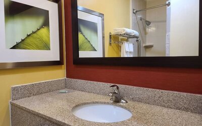 Hotel bathroom with granite vanity and sink, large framed mirror, yellow and red walls, decorative artwork and a shower with towels visible in the reflection.