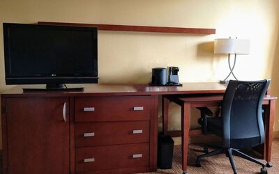 Hotel room workspace with wooden desk and storage unit, flat-screen TV, office chair, desk lamp, coffee maker and drawers against a warm yellow wall.