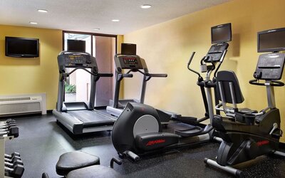 Hotel fitness center with treadmills, elliptical machines, stationary bike, dumbbells and workout bench on black rubber flooring with yellow walls.