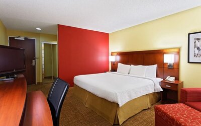 Warmly decorated hotel room with king bed, white bedding, wooden headboard, red accent wall, bedside lighting, red armchair and matching patterned ottoman.