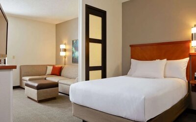 Spacious hotel day room with a king bed, separate sitting area with a sofa and ottoman, work desk, wall-mounted TV, large window, and warm contemporary décor.