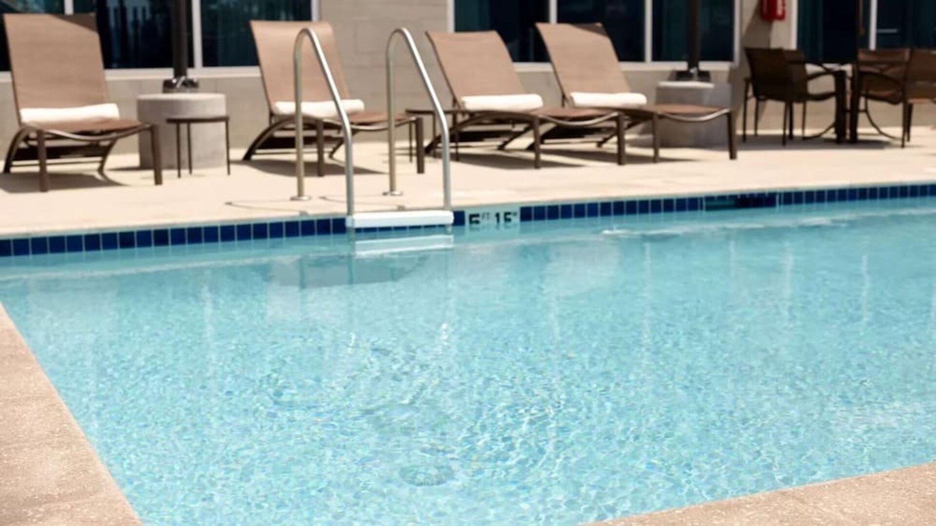 Hotel outdoor swimming pool with clear blue water, metal entry ladder, spacious pool deck, cushioned lounge chairs, side tables, and additional outdoor seating.