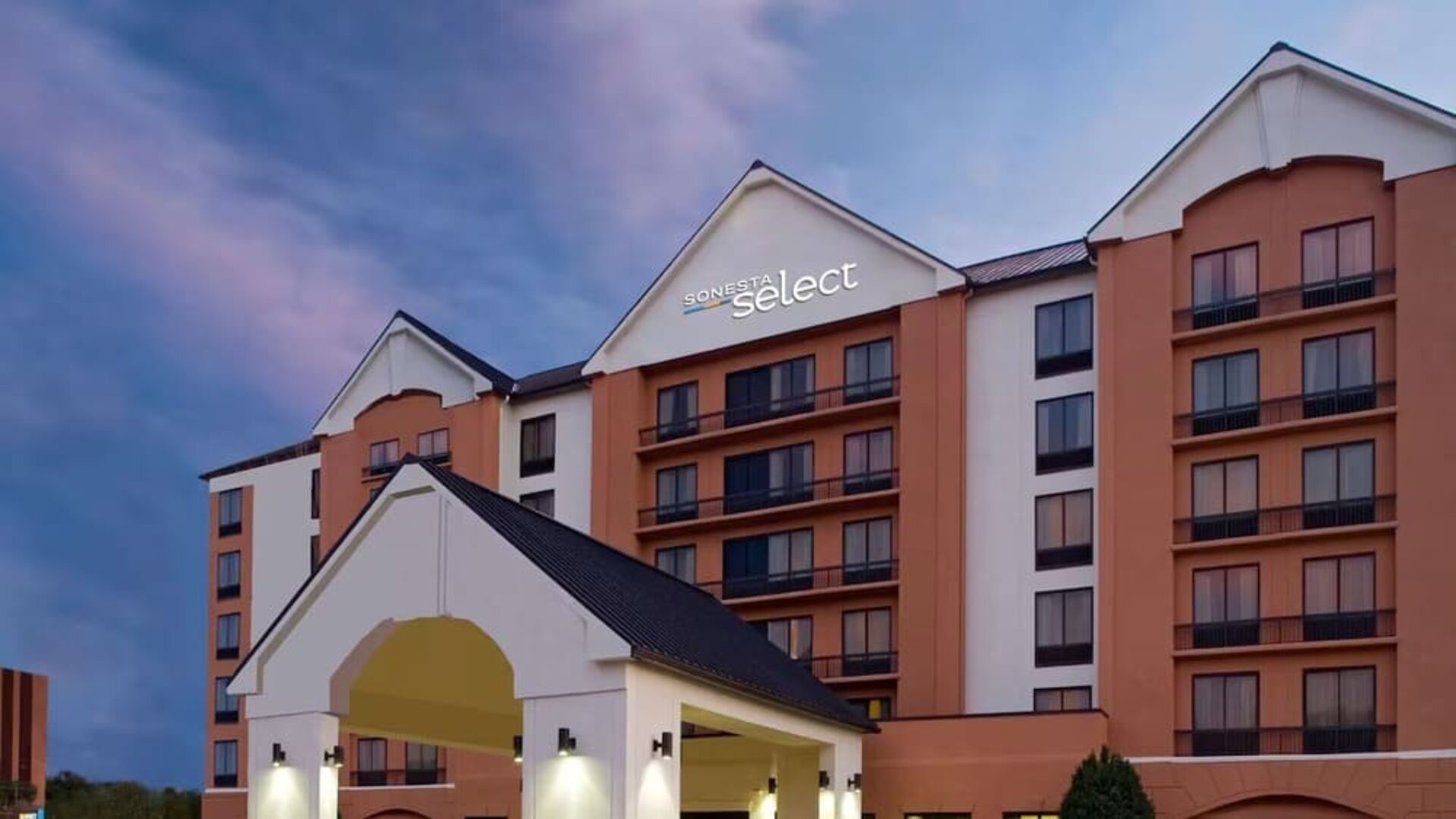 Exterior view of the multi-story Sonesta Select hotel at dusk, featuring a peach and cream façade, illuminated hotel signage, balconies, and a covered entrance.