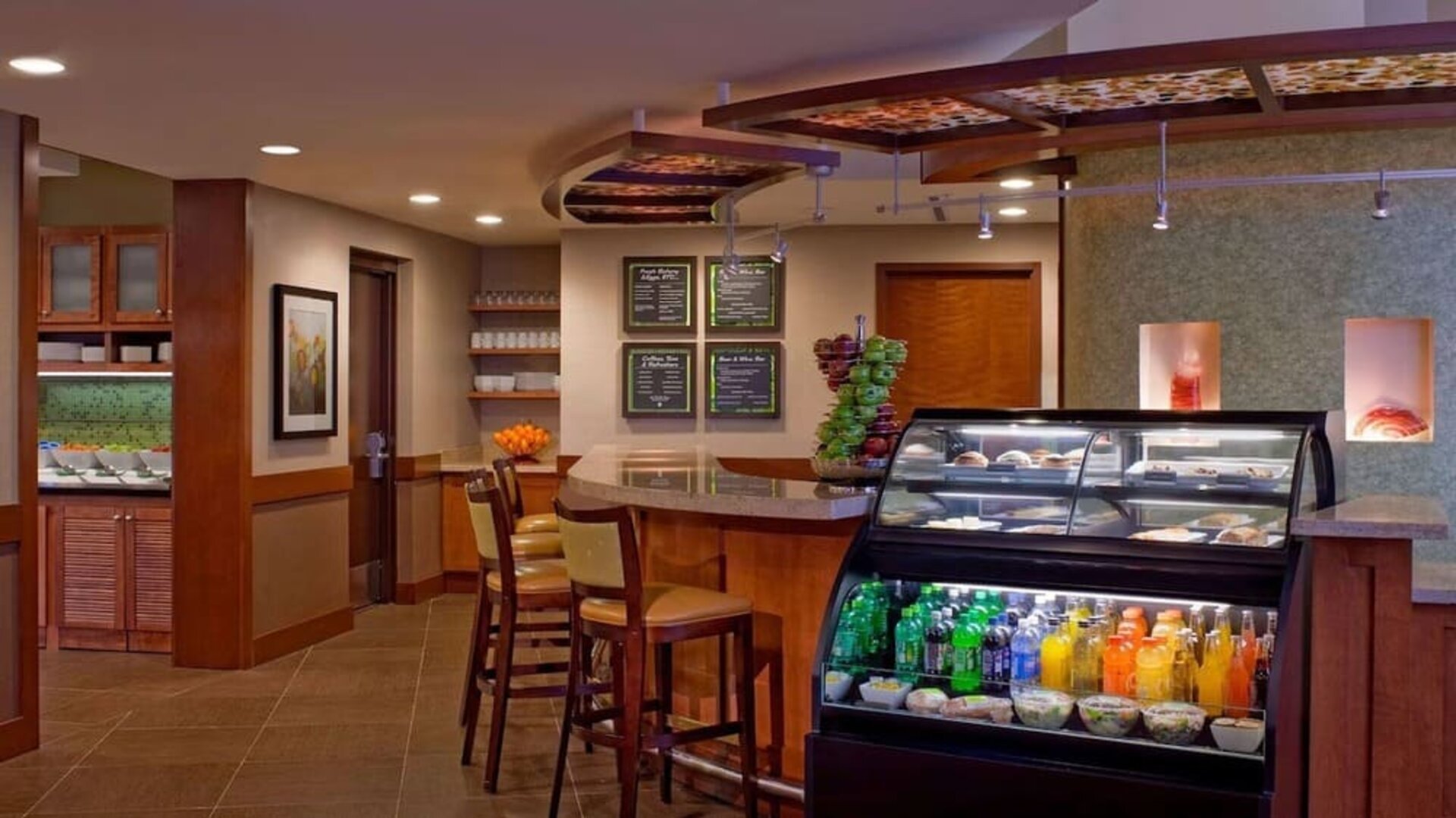 Casual hotel café and dining area with a refrigerated display case stocked with drinks and snacks, counter seating, wooden cabinetry, menu boards, and tables for guests.