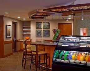 Casual hotel café and dining area with a refrigerated display case stocked with drinks and snacks, counter seating, wooden cabinetry, menu boards, and tables for guests.