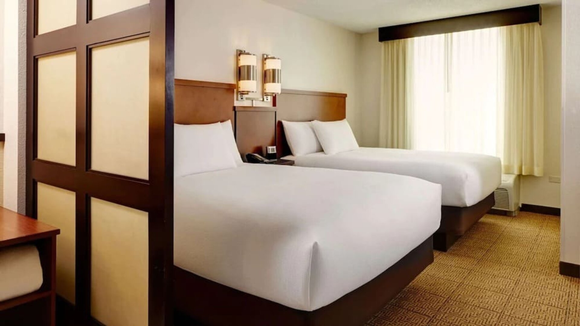 Spacious hotel room with two double beds dressed in white linens, wooden headboards, contemporary wall lights, a large curtained window, neutral carpeting, and a decorative room divider.