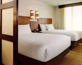 Spacious hotel room with two double beds dressed in white linens, wooden headboards, contemporary wall lights, a large curtained window, neutral carpeting, and a decorative room divider.