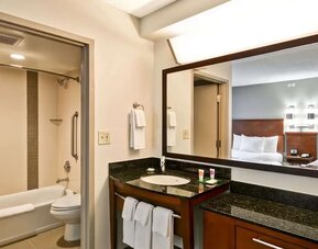 Modern hotel guest bathroom with a bathtub and shower, granite vanity with sink, large framed mirror, fresh white towels, and a view into a guest room with two beds.